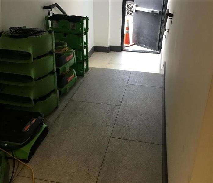 Seven SERVPRO air movers lined up against a hallway wall near an exit